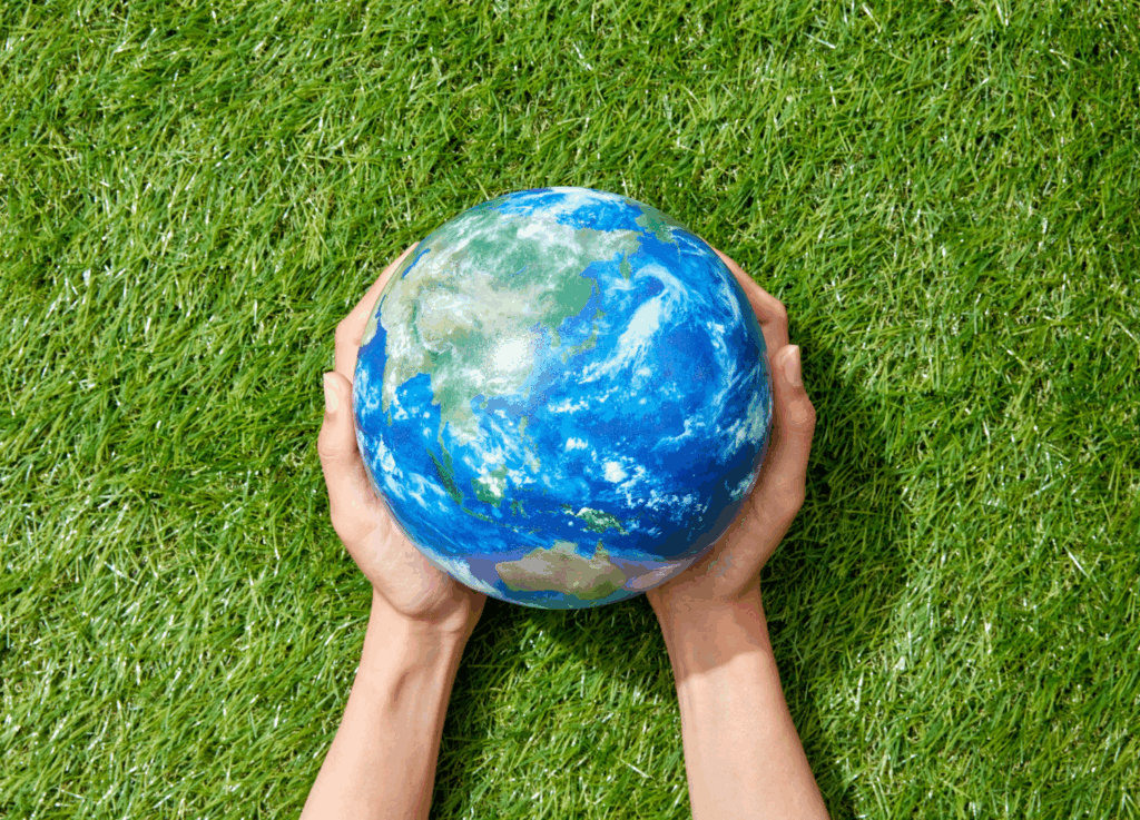 
Duas mãos seguram um globo terrestre, visto de cima, sobre um gramado verde. O globo é azul e branco, e representa os continentes e oceanos.