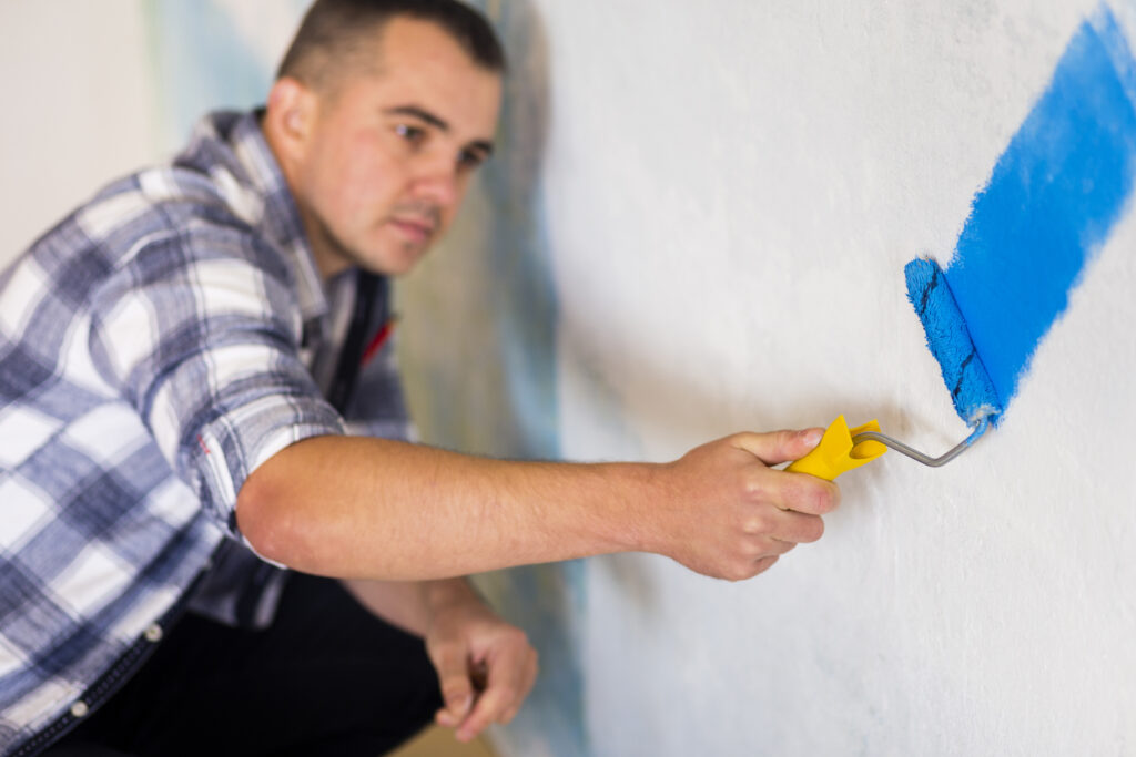 A imagem mostra um jovem construtor, de costas para a câmera, usando um capacete de segurança branco e um macacão azul por cima de uma camiseta laranja. Ele está pintando uma parede lisa, de cor laranja-clara, com um rolo de pintura.